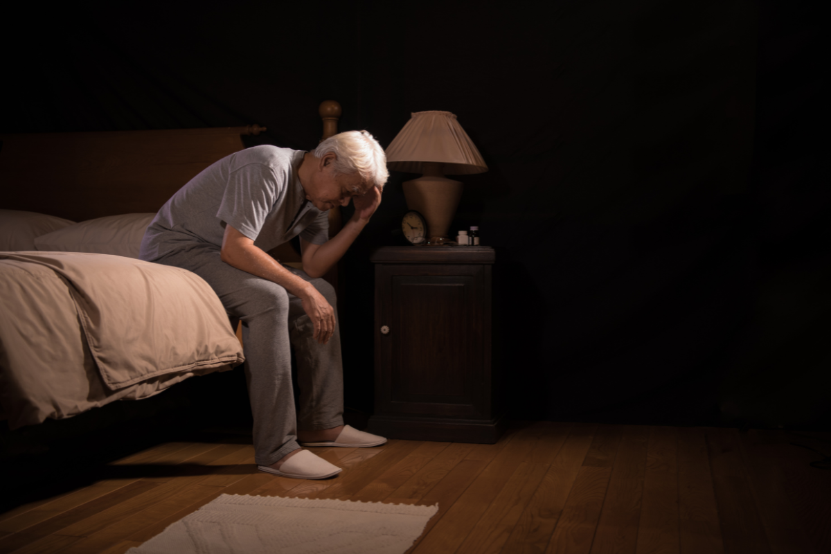 A man sitting on the edge of a bed in a dimly lit room, resting his head in his hand with a tired expression.