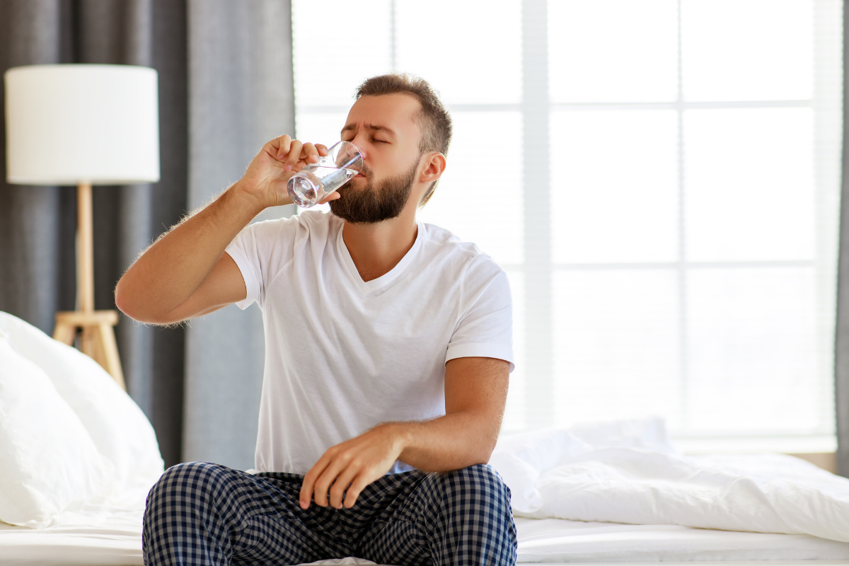 A person sits on a bed, drinking from a glass of water