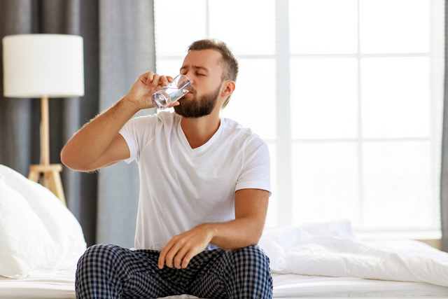 A person sits on a bed, drinking from a glass of water
