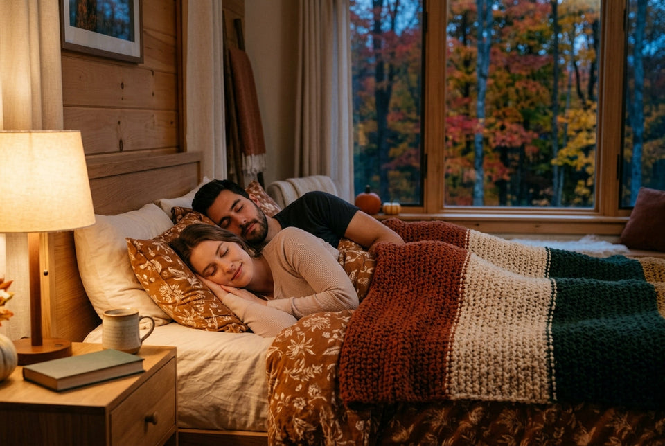 A couple sleeping peacefully under cozy knit blankets in a warm, rustic bedroom with large windows overlooking vibrant autumn foliage.